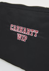 Pochette en tissu noir avec une texture lisse, arborant un logo "CARHARTT WIP" brodé en rouge. Fermeture éclair sur le dessus.