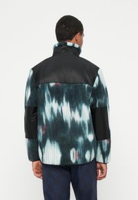 Fleece jacket with a black, padded top and a blue, tie-dye pattern. Features a high collar and textured fabric with contrasting black accents.