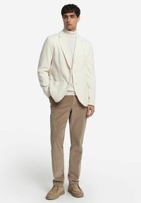 Leicht cremfarbener, strukturierten Blazer über einem weißen Rollkragenpullover, kombiniert mit beigen Cordhosen und taupefarbenen Slippern.