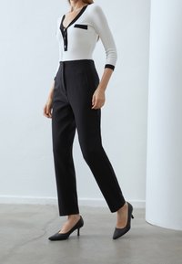 Top bianco a maniche lunghe con dettagli neri e bottoni, abbinato a pantaloni neri a vita alta e tacchi alti neri, su una superficie in cemento liscio.