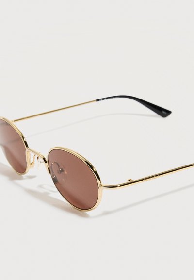 Στρογγυλά γυαλιά ηλίου με λεπτό χρυσό μεταλλικό σκελετό και φακούς με καφέ απόχρωση, τοποθετημένα σε λευκή επιφάνεια.