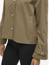 Olive green jacket with a structured design, featuring large buttons, a side pocket, and accentuated cuff details. Paired with black pants.