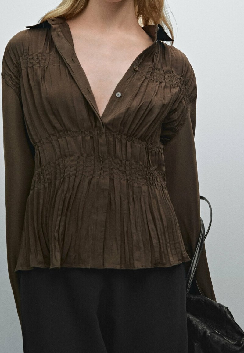 Femme portant un chemisier marron plissé avec des détails smockés, un pantalon noir, et tenant la lanière d'un sac à main en cuir noir sur l'épaule.