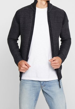 Pull noir en maille à fermeture éclair avec une texture côtelée et un col montant, porté sur un t-shirt blanc, associé à un jean en denim bleu.