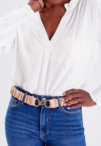Person wearing a white V-neck blouse and blue jeans with a beige ruched belt fastened by a gold circular clasp at the waist.