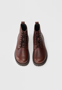 Par de botas de piel marrón hasta el tobillo con cordones en la parte frontal y suelas de goma negra, mostradas desde arriba sobre un fondo blanco.