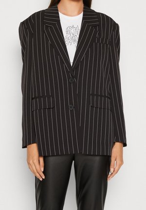 Kvinde iført en sort dobbeltradet blazer med nålestriber og hakkede revers over en hvid t-shirt med grafik og sorte læderbukser.