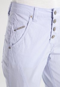 Ljuslavendelfärgade byxor med en sidficka som har en metalltagg märkt "OPUS," med fem knappdetaljer längs midjan.