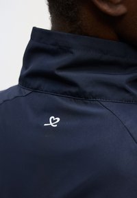 Πλάτη ενός ατόμου που φοράει σκούρο μπλε μπουφάν με ψηλό γιακά και μικρό λευκό λογότυπο σε σχήμα καρδιάς στην πάνω πλάτη.