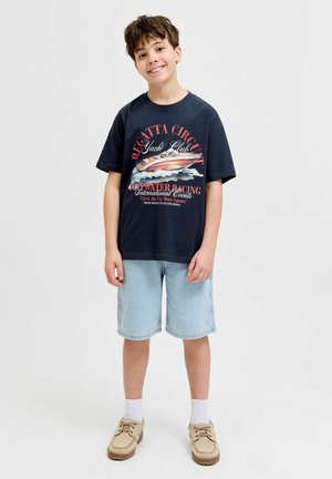 Usmievajúci sa chlapec stojí oblečený v čiernom tričku s potlačou jachtového klubu, svetlých džínsových kraťasoch, bielych ponožkách a béžových šnurovacích topánkach na jednofarebnom pozadí.