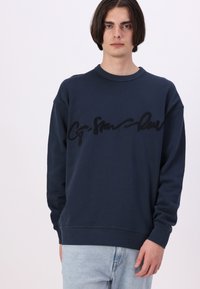 Námornícky modrý sweatshir s okrúhlym výstrihom a dlhými rukávmi, s výrazným čiernym vyšitým logom na hrudi. Mäkká textura.