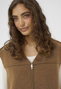 Smeđi vuneni prsluk s patentnim zatvaračem sprijeda, koji ima suptilnu teksturu i visok izrez. Nosi se preko svijetle majice.