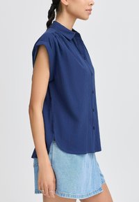 Camicia blu navy a maniche corte con bottoni, realizzata in un tessuto testurizzato, con colletto a punta e spacchi laterali, abbinata a una gonna di jeans azzurri.