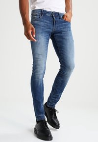 Hombre vistiendo jeans azul ajustados y zapatos de vestir negros, de pie con una mano en el bolsillo frente a un fondo liso.