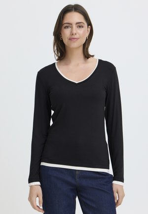 Žena s hnedými vlasmi po ramená, oblečená v čiernom dlhorukávovom tričku s výstrihom do V a bielym lemovaním, modrých džínsoch, stojaca pred jednoduchým pozadím.