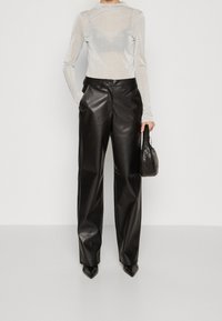 Person med en genomskinlig silverfärgad långärmad topp, svarta läderbyxor med vida ben, spetsiga svarta skor, som håller en svart kviltad handväska.