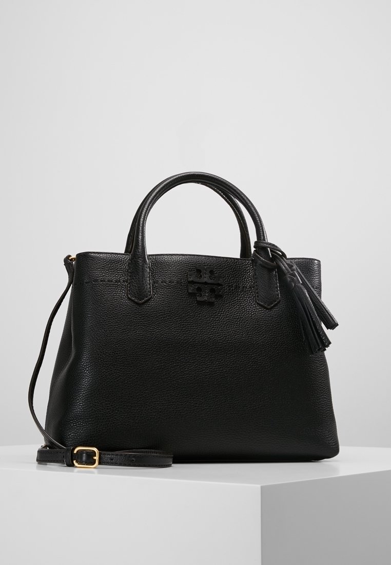 Sac à main en cuir noir texturé avec doubles poignées, pompon fixé, logo à l'avant et bandoulière ajustable avec boucle dorée.