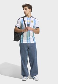 Mladý muž v argentinském fotbalovém dresu, s širokými džínami, bílými teniskami a černou taškou přes rameno, dívající se na stranu.