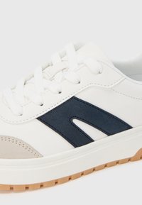 Balta sportinė bateliai su tamsiai mėlynu šoniniu akcentu, tekstūruotu viršumi, plokščiais baltais raišteliais ir šviesiai rudos gumos padu.