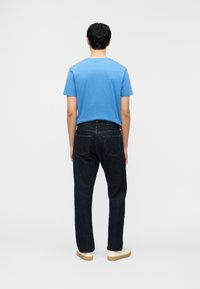 Άνδρας που στέκεται με την πλάτη γυρισμένη, φοράει μπλε κοντομάνικο πουκάμισο, σκούρο τζιν και λευκά αθλητικά παπούτσια σε ένα απλό φωτεινό φόντο.