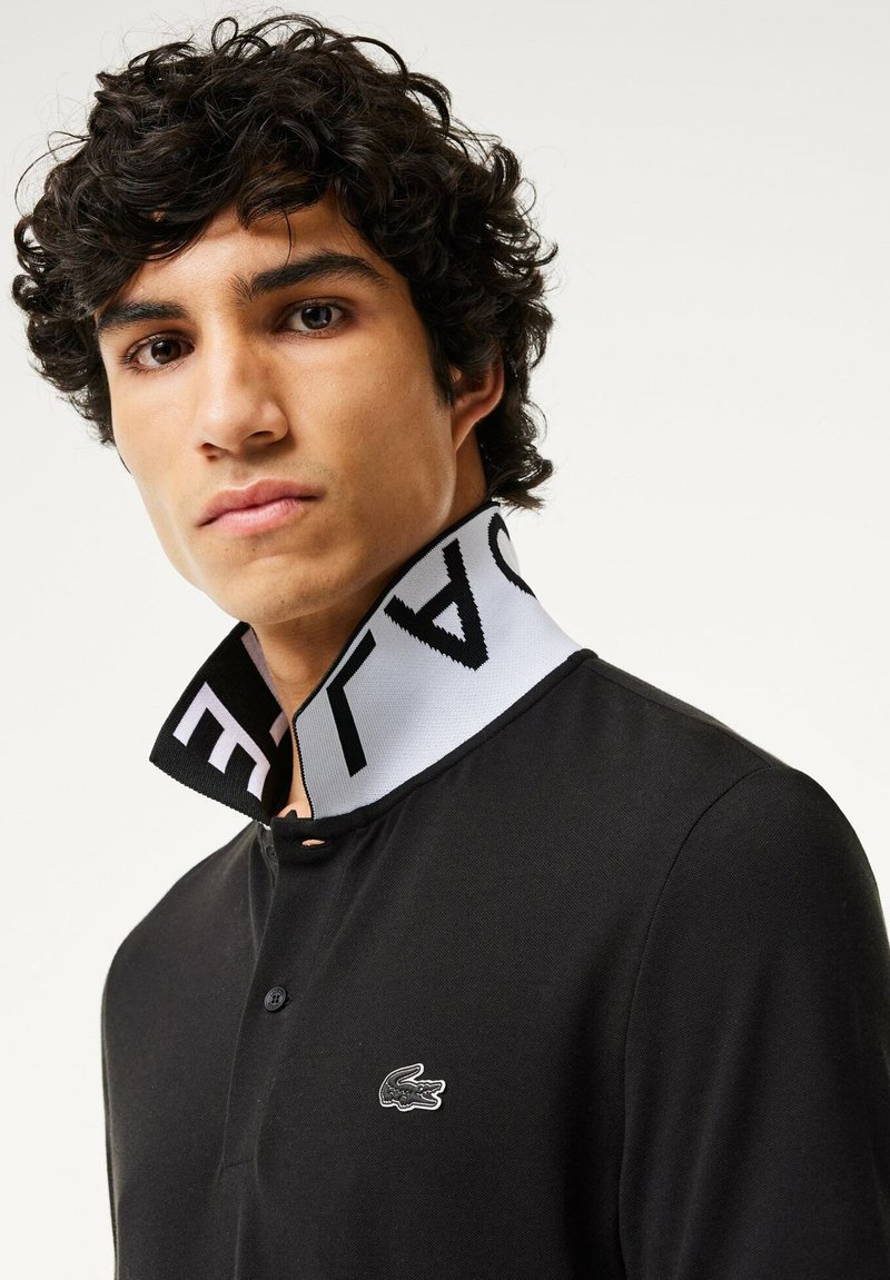 Schwarzes Polohemd mit spitzem Kragen und einem kontrastierenden weißen Band, das "LACOSTE" zeigt. Kleines Logo auf der Brust. Glatte Textur.