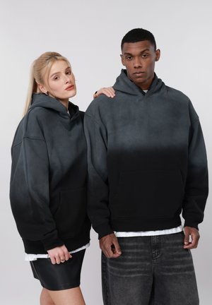 Dvaja mladí dospelí v identických čiernych ombré mikinách stoja vedľa seba, jeden si položil ruku na rameno druhého, na jednoduchom pozadí.