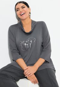 Top gris con cuello de cowl y mangas tres cuartos, con bordado floral blanco. Combinado con pantalones anchos de pierna amplia en gris oscuro.