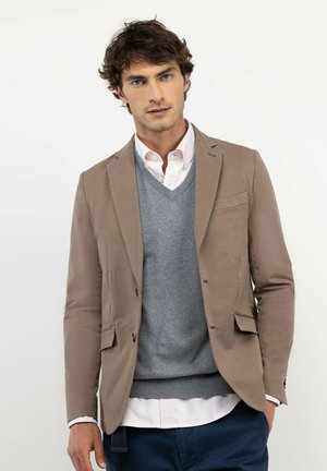Hombre con blazer beige, suéter gris con cuello en V, camisa blanca y pantalones azul oscuro sobre un fondo claro y liso.