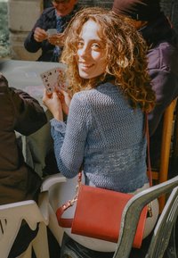 Röd lädercrossbody-väska med guldaccent; ljusblå stickad tröja med intrikat mönster; sitter vid ett bord med spelkort.