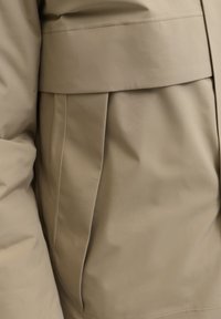 Beige Oberbekleidung mit glatter Textur, strukturierter Form und plissierten Details an der Seite. Gestylt mit einem breiten Gürtel in der Taille.