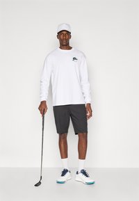 Fehér, hosszú ujjú ing zöld logóval, fekete nadrág, fehér golfcipő kék akcentussal, és fehér sapka; egy golfütőt tart.