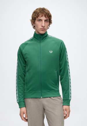 Junger Mann trägt eine grüne Jacke mit Reißverschluss und weißen Logos auf den Ärmeln und der Brust, kombiniert mit hellgrauen Hosen, steht vor weißem Hintergrund.