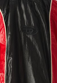 Sort og rød jakke med en glat, skinnende overflade. Har kontrasterende hvide detaljer og et broderet logo på det sorte stof.