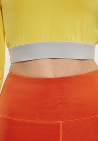 Geel crop top met een grijze elastische tailleband, gecombineerd met hooggeknoopte oranje legging, met gladde texturen en stiksels.