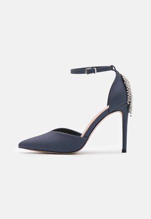 Granatowy teksturowany but na wysokim obcasie z ostrym noskiem, paskiem wokół kostki z klamrą oraz zdobieniem z kryształków na tyle buta.