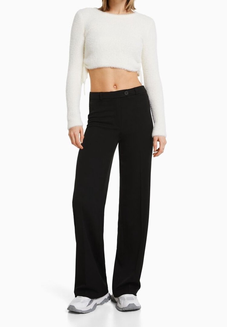 Femme portant un pull court blanc à manches longues, un pantalon noir à jambes larges et des baskets blanches, debout devant un fond uni.