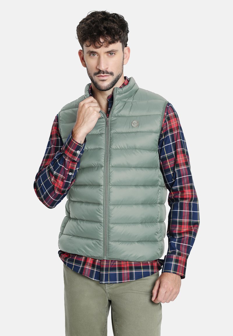 Chaleco verde acolchado con cierre de cremallera y cuello alto, llevado sobre una camisa de cuadros roja y azul. Presenta un logo en la parte superior izquierda.