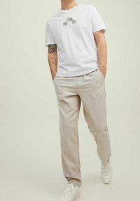 Biała bawełniana koszulka z grafiką palmy, zestawiona z jasnobeżowymi lnianymi spodniami i białymi trampkami. Model stoi z rękami w kieszeniach.