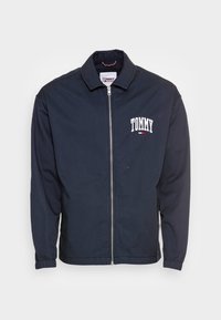Marinblå bomberjacka med full dragkedja, ståkrage och elastiska muddar. Har en vit broderad "TOMMY"-logotyp på vänster bröst.
