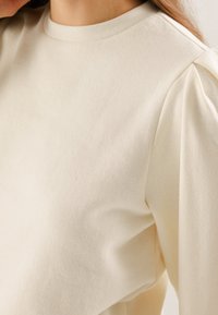 Krémový pulover s kulatým výstřihem, texturovaná tkanina a nashromážděné balónové rukávy pro přidanou hloubku. Hladké, jemné detaily designu.