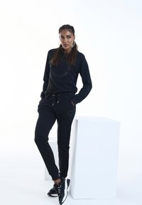 Set di felpa e pantaloni neri realizzato in tessuto morbido, con un logo rotondo in verde scuro. Include un elastico in vita con cordino e tasche laterali.