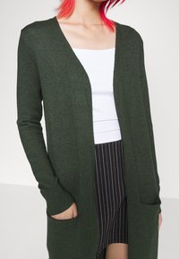 Mörkgrön kofta med V-ringning, långa ärmar, ribbad struktur och två frontfickor. Vit undertröja och rutigt nederdel synlig.