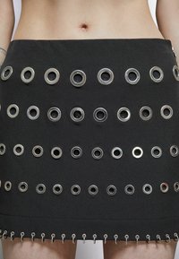 Jupe mini en tissu noir avec plusieurs œillets en métal argenté disposés en rangées, avec un bord inférieur froncé et une fermeture éclair visible.
