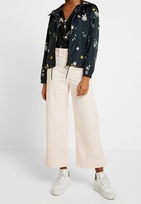 Czarna kurtka przeciwdeszczowa w kwiaty z zielonymi i żółtymi akcentami, w połączeniu z szerokimi kremowymi spodniami i białymi trampkami.