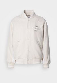 Biela fleecová bunda s rebrovaným golierom, bočnými pruhmi a predným zapínaním na gombíky. Obsahuje vyšívaný nápis "Tommy Jeans" v tučnom písme.