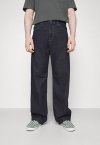 Ciemnoniebieskie jeansy z denimu o prostych nogawkach, zapinane na górny guzik, z żółtym przeszyciem, zestawione z zielono-białymi wsuwanymi butami w kratkę.