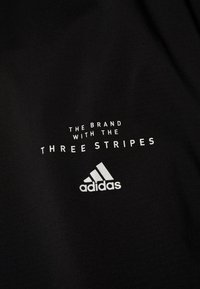 Svart tyg med vit grafisk text som säger "MÄRKET MED DE TRE RANDERNA" och Adidas-logotypen i nedre högra hörnet.