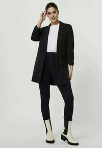 Um casaco longo preto sobre uma t-shirt branca, combinado com jeans escuros e botas de tornozelo creme. O casaco possui um design elegante e ajustado.