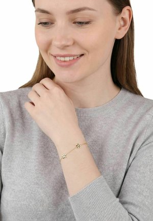 Młoda kobieta w szarym swetrze uśmiecha się, pokazując na nadgarstku złotą bransoletkę z dwoma zawieszkami w kształcie kwiatów.