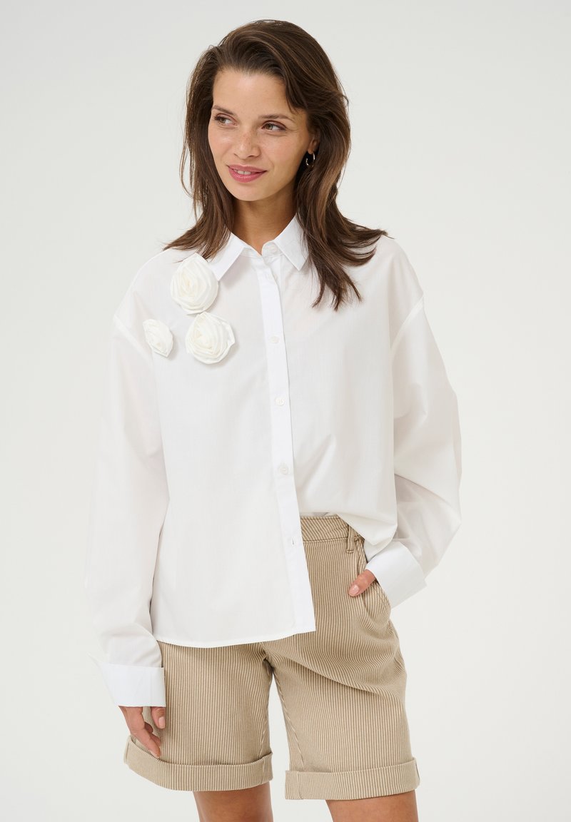 White button-up shirt with three white fabric flower accents on the left. Paired with beige striped shorts, showing a relaxed fit.